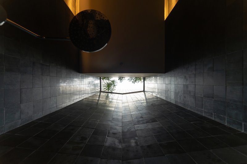 Covered entrance passage with dark stone floor and slatted walls leading to a sunlit courtyard with greenery