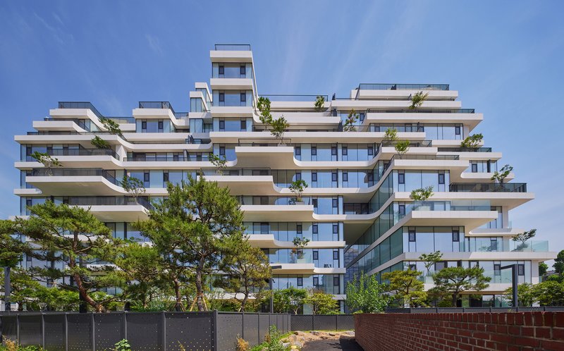 Terraced residential building with staggered white balconies and floor-to-ceiling glass behind mature pines