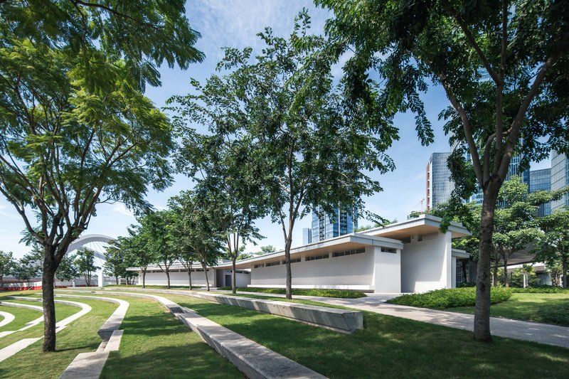 Low-slung roof volume extending through a park with curved pathways and mature canopy trees