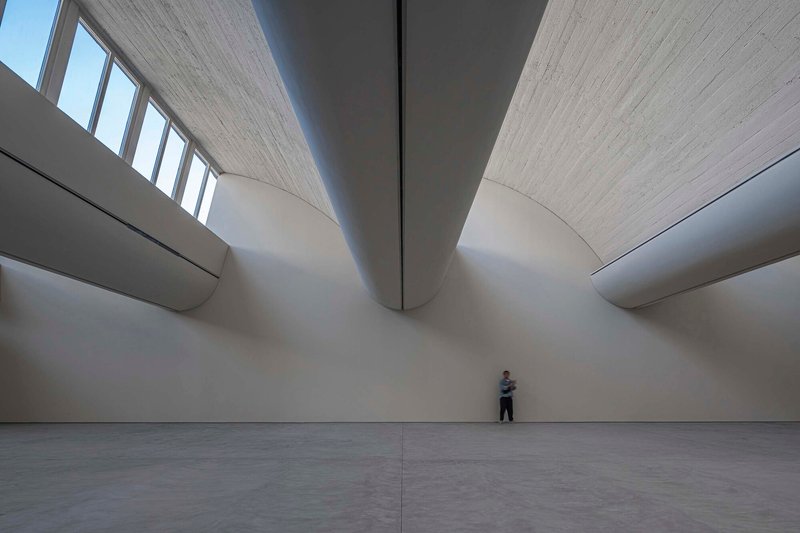 Interior gallery space with vaulted timber ceiling and clerestory windows above curved white walls with a visitor