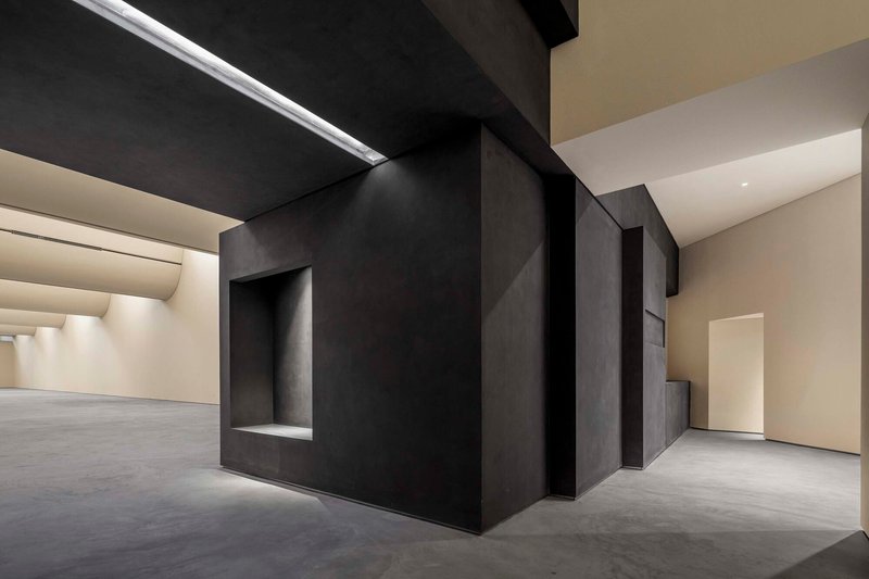 Dark metallic exhibition wall with recessed niche alongside white gallery spaces under artificial light