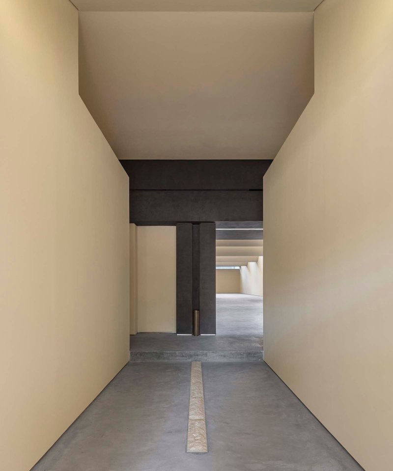 Narrow passage with limestone floor inlay leading through dark concrete frame into stepped ceiling gallery