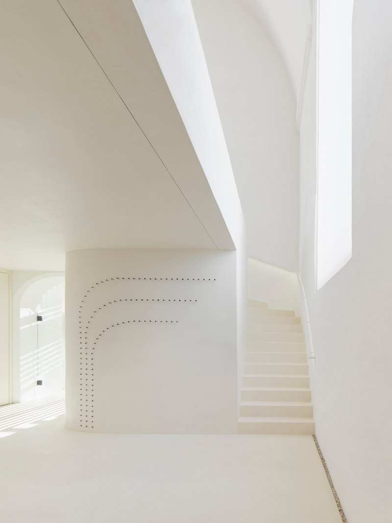 Curved white staircase ascending alongside a perforated wall beneath a skylight slit