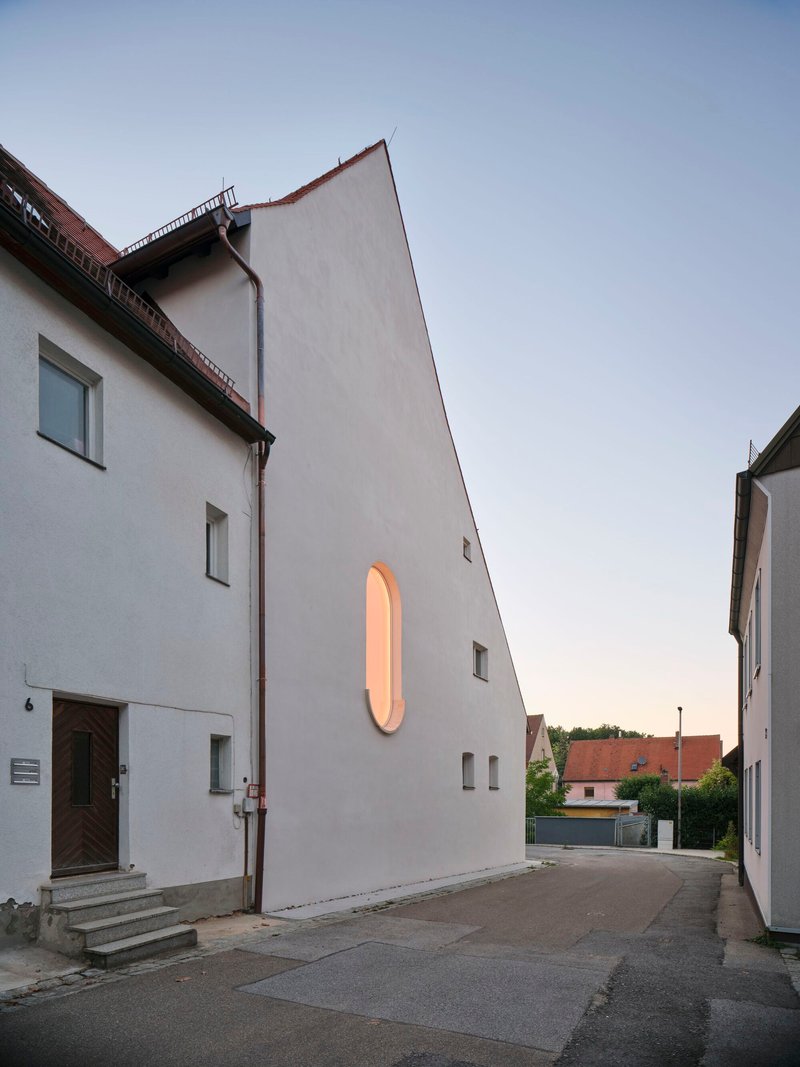 White rendered facade with an illuminated oval window opening facing a quiet street at dusk