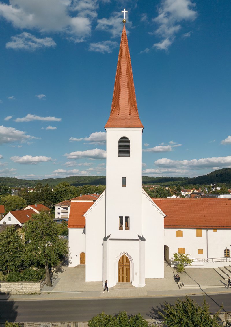 Front elevation of a white church with terracotta spire and timber arched door under a bright blue sky