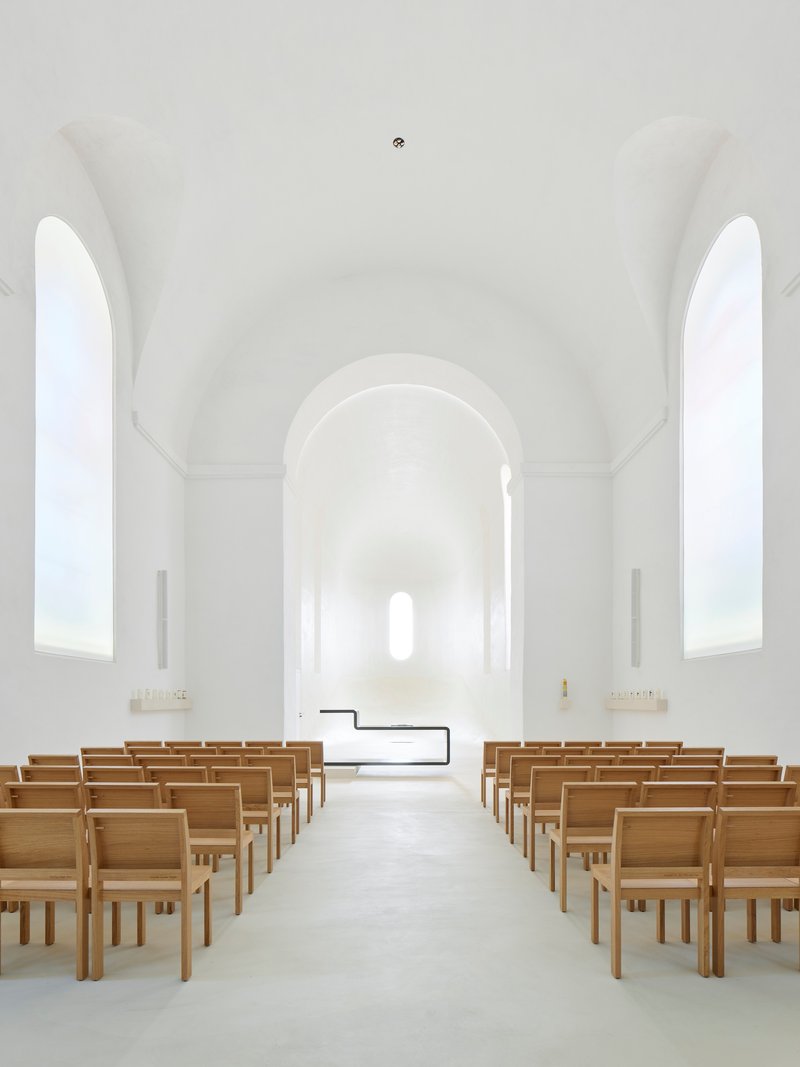 White vaulted chapel interior with timber pews flanking a central aisle leading to arched apse