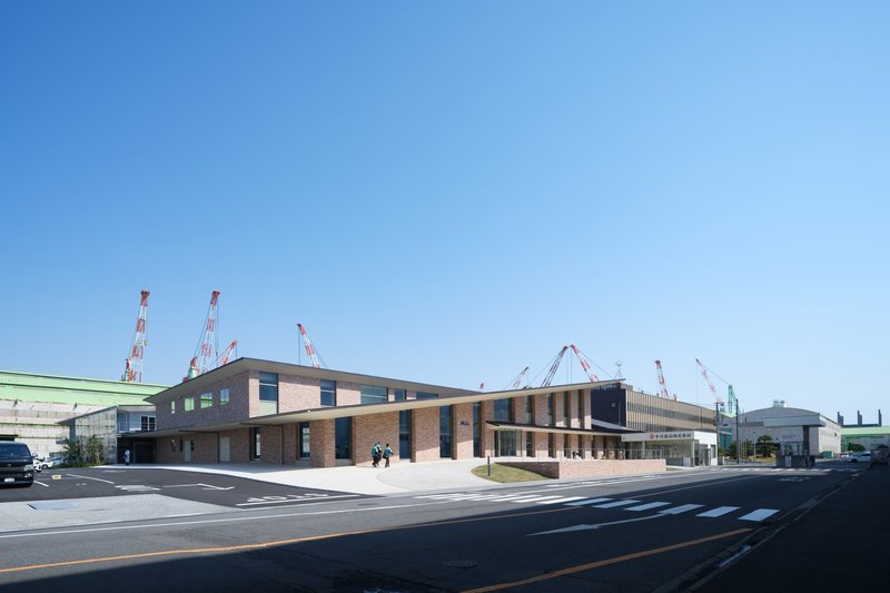 Long facade showing alternating brick volumes with vertical sunshades and pedestrians crossing at the entrance