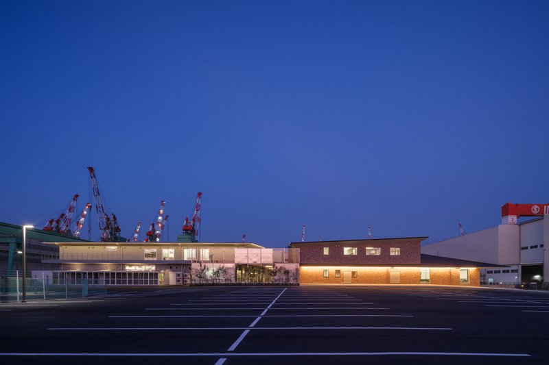 Illuminated one and two-story volumes across an empty parking lot with shipyard cranes in deep blue hour