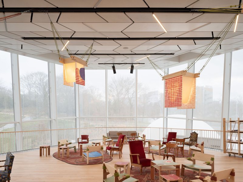 Glass-walled upper floor activity room with geometric ceiling, suspended fabric screens, and misty trees beyond
