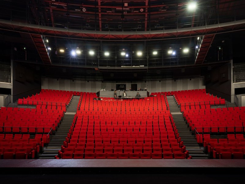 Theater auditorium with tiered red seating, exposed steel trusses, and stage-level lighting