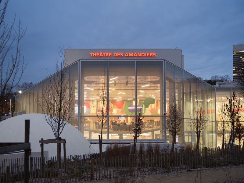 Glass facade of the theatre building illuminated at dusk with bare winter trees in the foreground