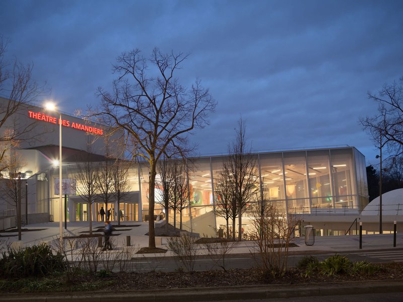 Street view of the illuminated glass-wrapped lobby and entrance plaza framed by bare trees at twilight
