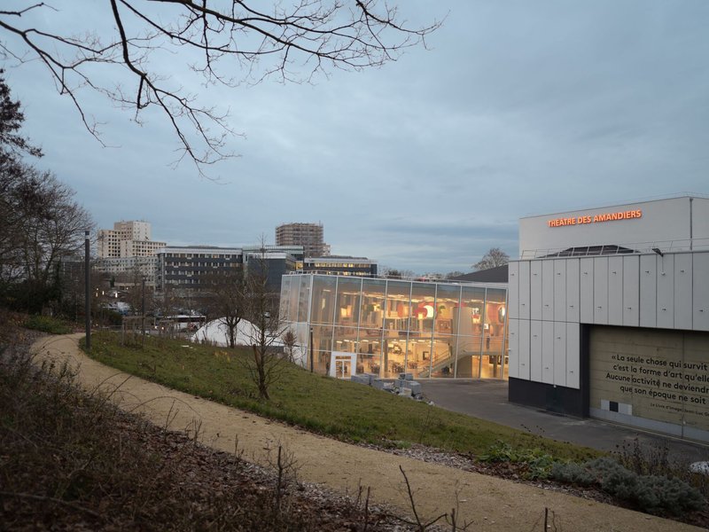 Illuminated glass facade with metal panel cladding viewed from a park path at dusk