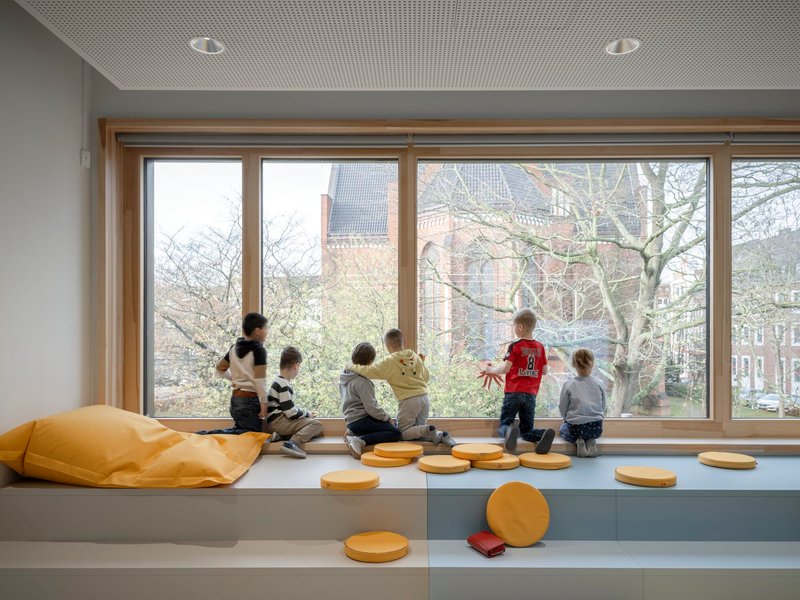 Window bench with children seated facing bare branches and brick buildings outside