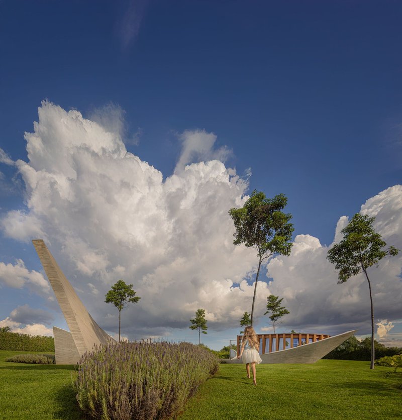 Curved concrete walls and timber colonnade framing the lawn under towering cumulonimbus clouds