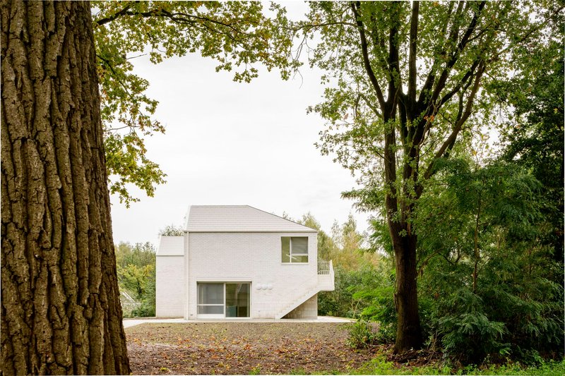 Rear elevation of white brick volumes with gabled roofs seen through mature deciduous trees