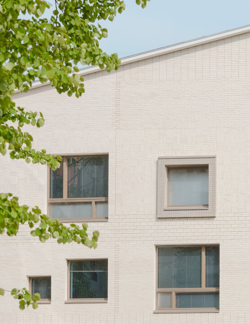 Pale brick facade with scattered windows framed by green tree branches under overcast sky