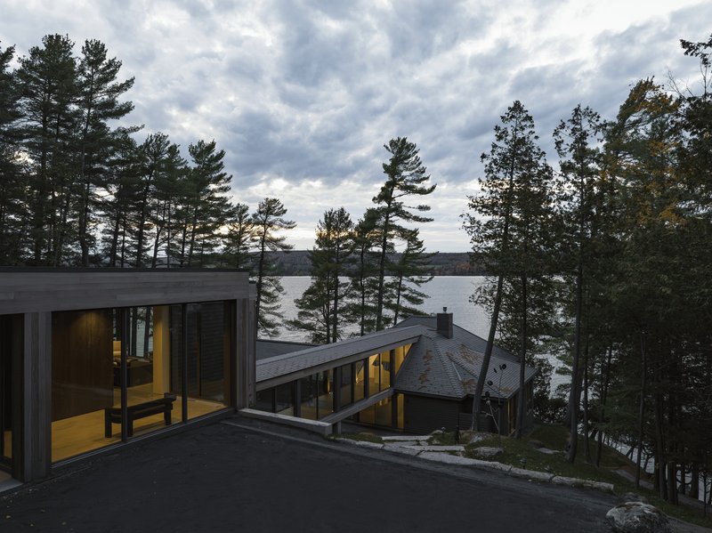 Exterior view of dark-clad volumes among pines overlooking a lake at dusk