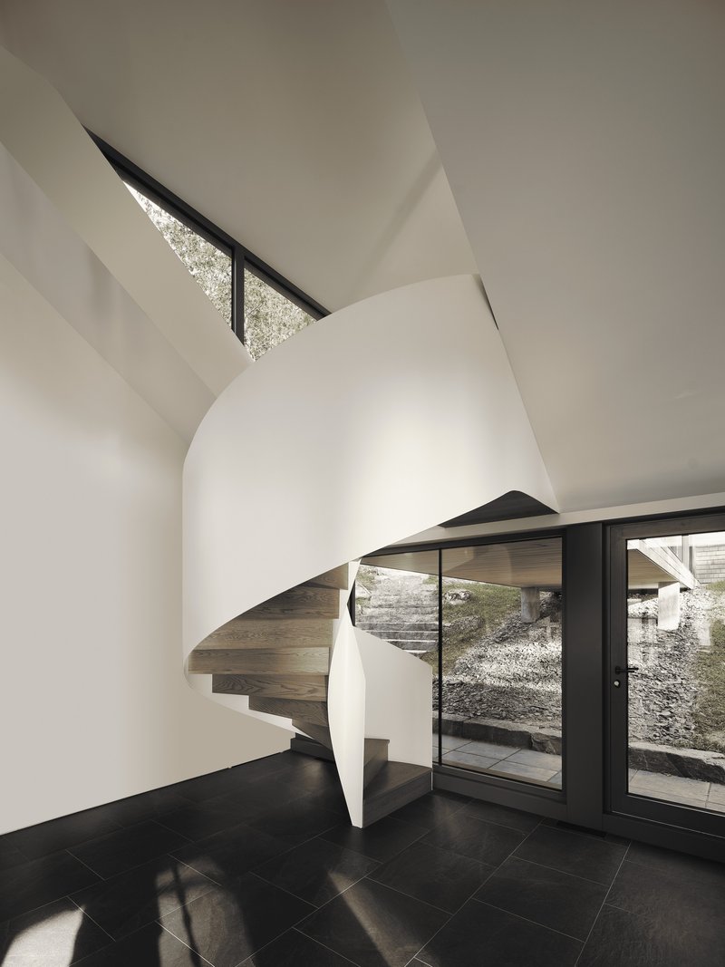 Curved sculptural staircase wrapped in white walls beside dark-framed glass doors opening to courtyard