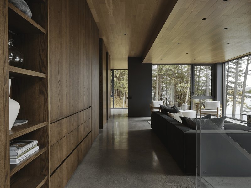 Living room with walnut built-in shelving, polished concrete floor, and floor-to-ceiling windows overlooking a winter forest