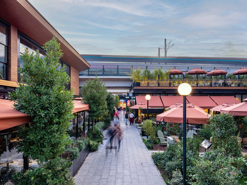Pedestrian promenade lined with planters and cafe umbrellas beneath the elevated rail line at dusk