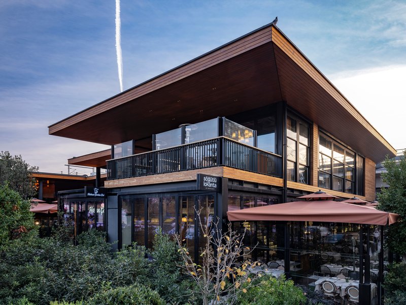 Two-story pavilion with deep overhanging timber eaves and terracotta awnings amid landscaped plaza at twilight