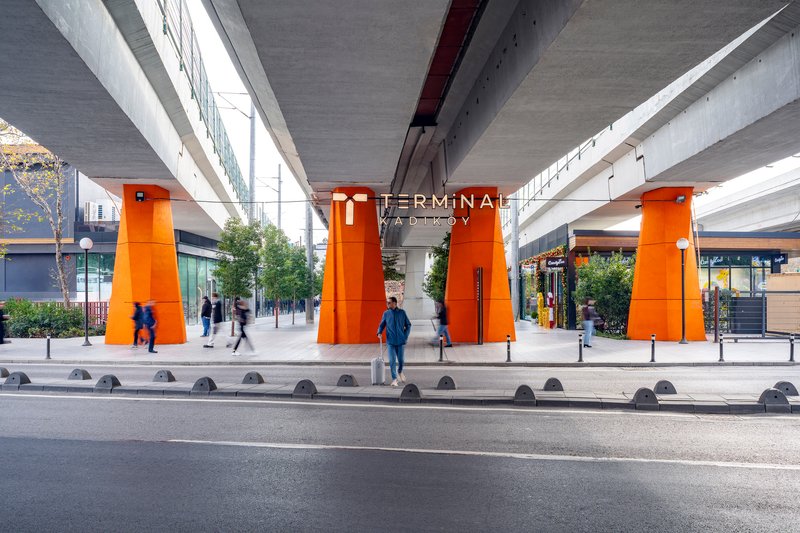 Street view of orange-clad steel columns supporting concrete viaduct with pedestrians crossing at grade