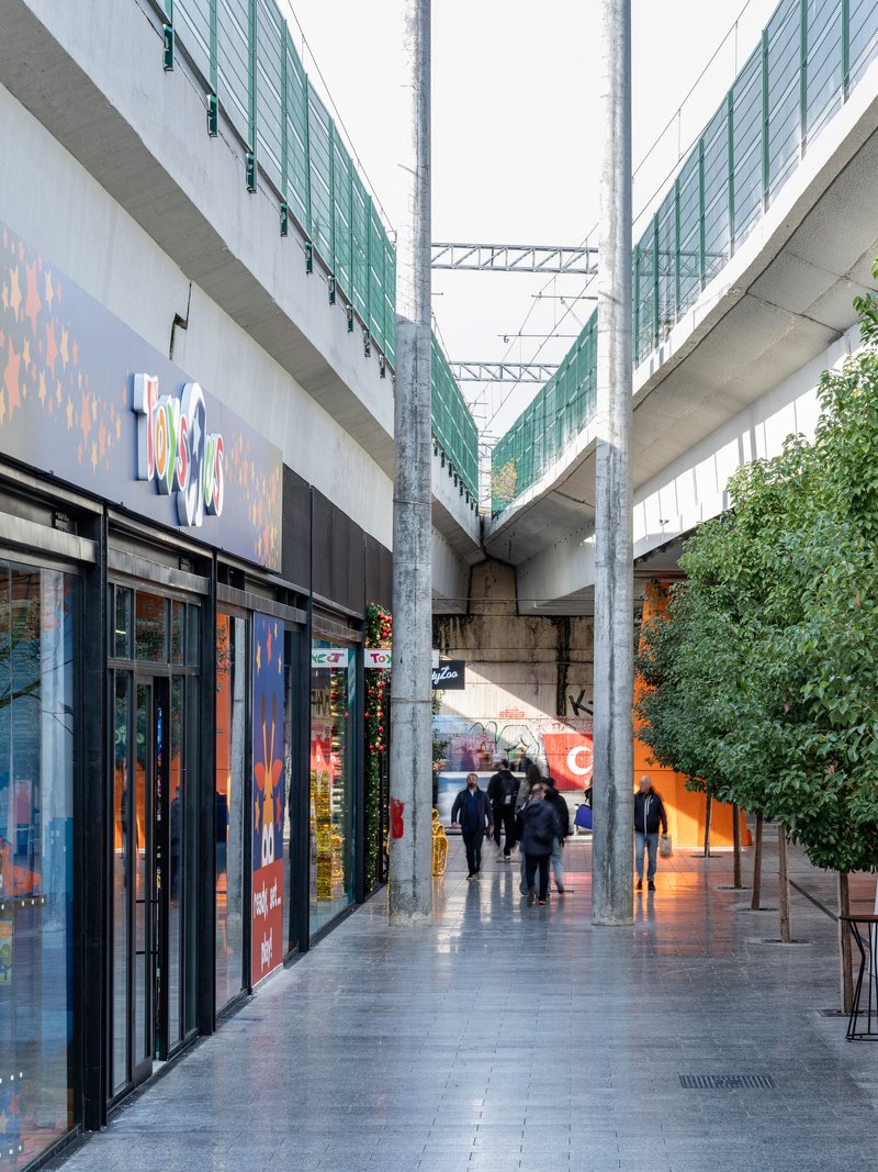 Glazed corridor beneath the concrete viaduct with retail storefronts and pedestrians on wet pavement