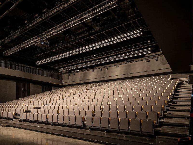 Auditorium with raked timber seating benches, exposed metal truss ceiling, and theatrical lighting rig overhead