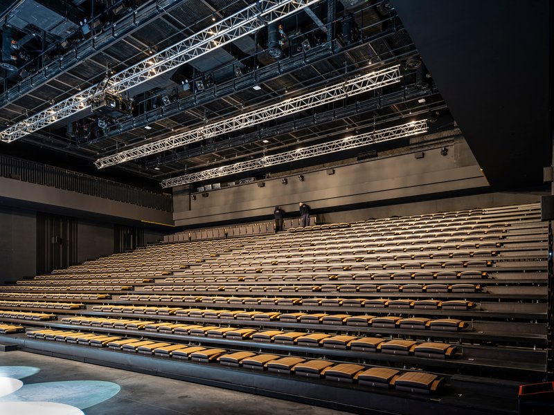Auditorium seating with two figures standing at the upper tier of wooden benches under theatrical lighting