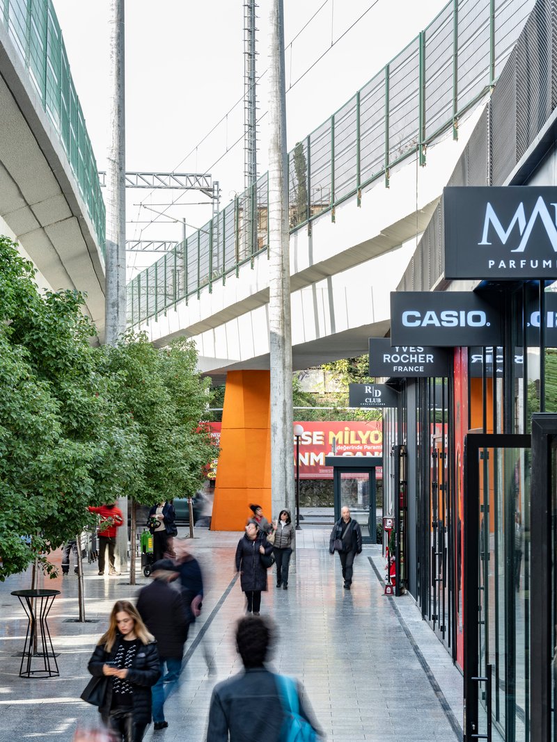 Shopping passage with polycarbonate screen walls and planted trees between retail facades at dusk