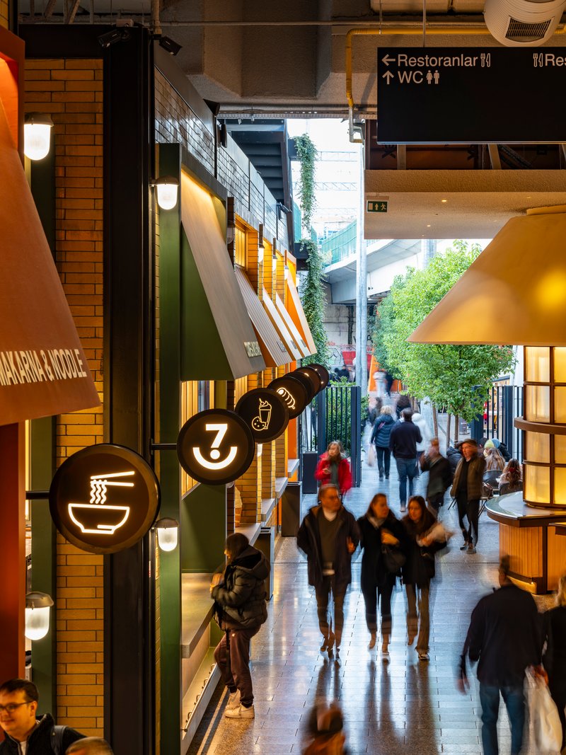 Food hall corridor with angled awnings and circular signage as visitors walk toward daylit courtyard