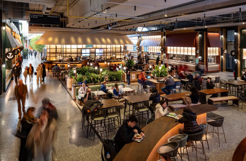 Central food hall seating area with communal tables beneath illuminated kiosk canopies and exposed ceiling systems