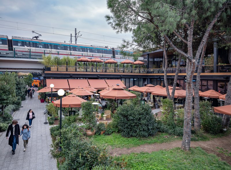 Open courtyard with terracotta umbrellas and pedestrians walking beneath an elevated train passing overhead