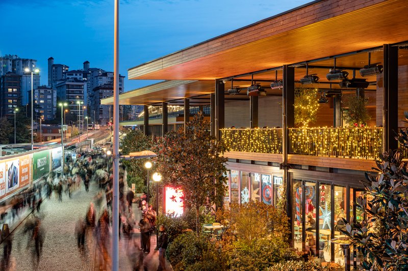 Timber-lined canopy extending over a two-story glass facade with string lights and evening crowd below