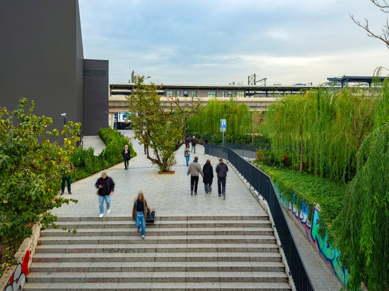 Stone staircase descending to a planted pedestrian plaza with people walking under cloudy skies