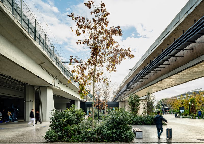 Concrete transit viaducts flanking a planted courtyard with a person walking past shrubs in afternoon light