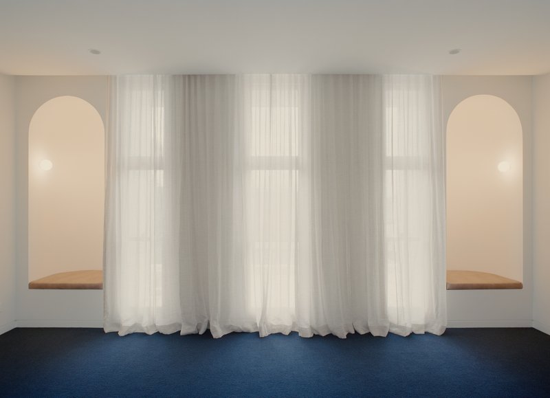 Symmetrical wall with twin arched niches and timber window seats behind floor-length sheer curtains over blue carpet