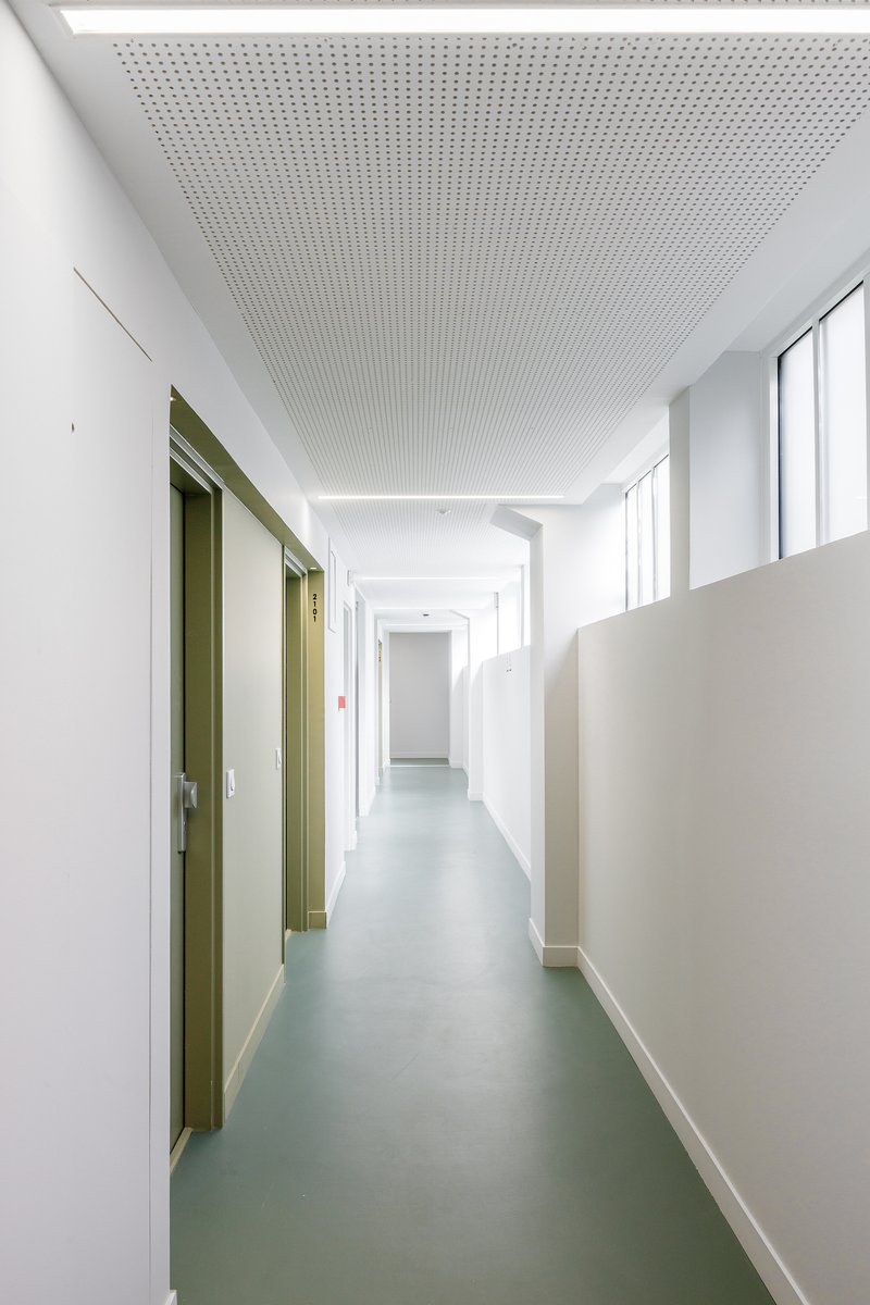 Corridor with perforated acoustic ceiling panels and green rubber flooring leading to olive-green unit doors