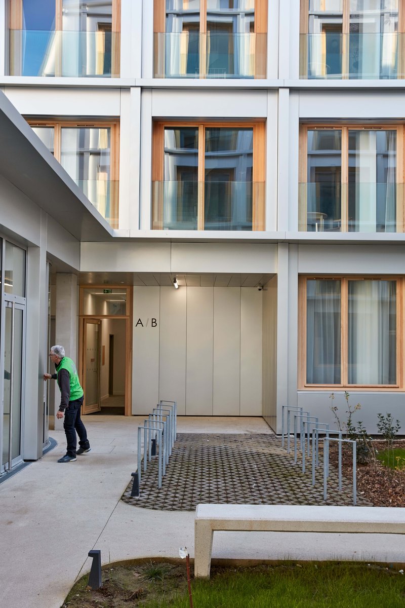 Courtyard entrance with person in green jacket walking toward timber doors and planted bed