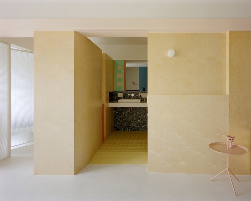 View through pale plywood volumes toward a bathroom with terrazzo vanity and yellow tile floor