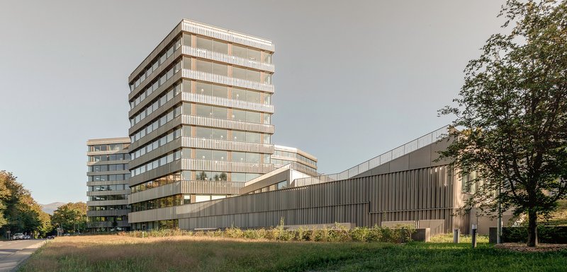 Facade composition with vertical louvered tower and horizontal ribbed base across a wildflower meadow