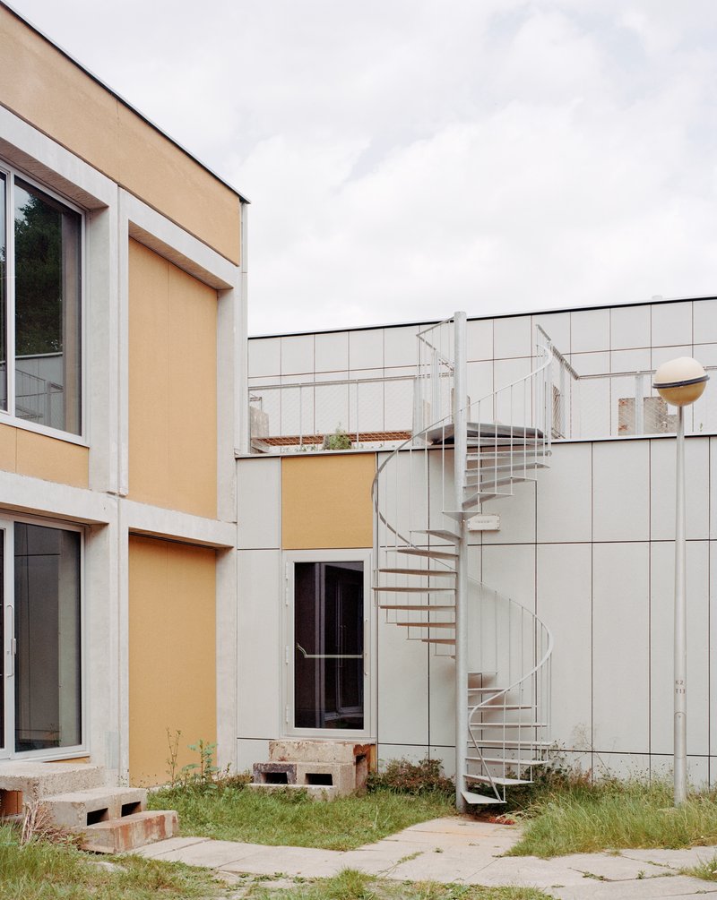 Exterior white spiral stair against white tile cladding with grassy courtyard and orange panel volumes