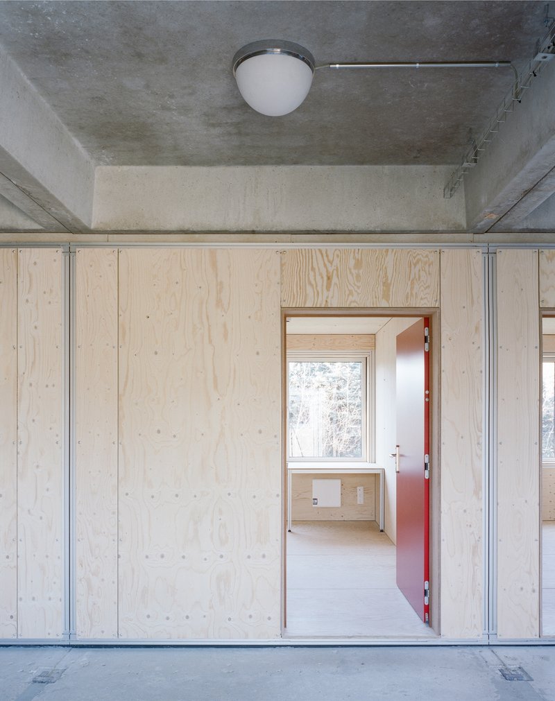 Light plywood wall with salmon-colored door opening to room with exposed concrete ceiling and steel channels
