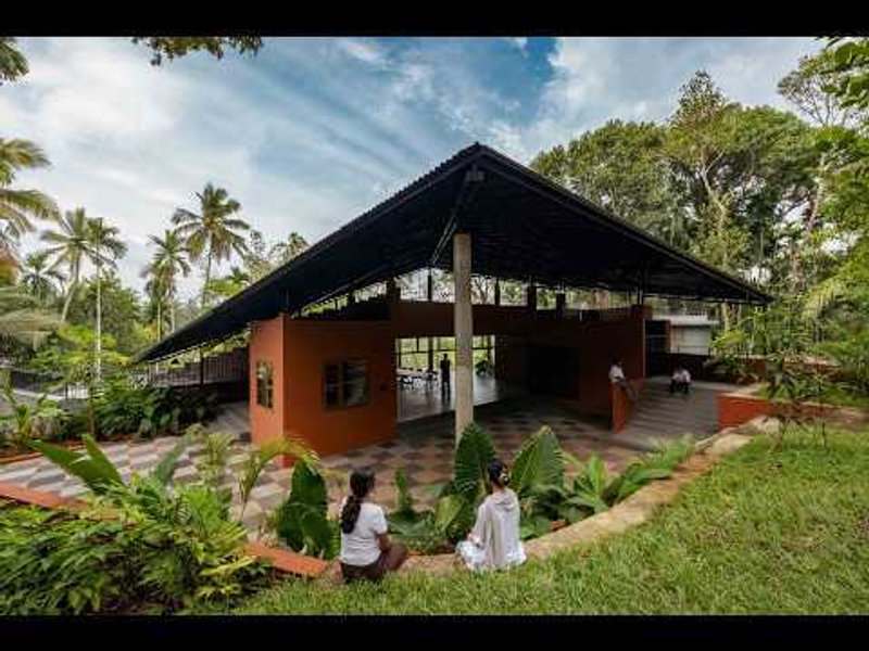Covered pavilion with terracotta walls and dark sloping roof surrounded by lawn and palm trees