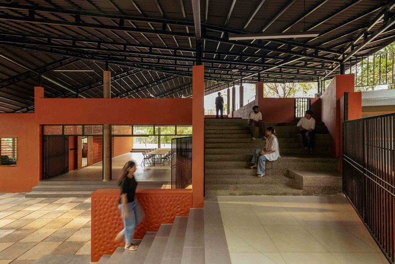 Interior hall with exposed steel roof trusses, terracotta walls and concrete staircase with people seated