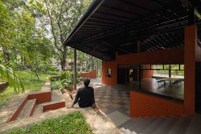 Covered terrace with person seated beside planted bed looking toward interior communal space and surrounding forest