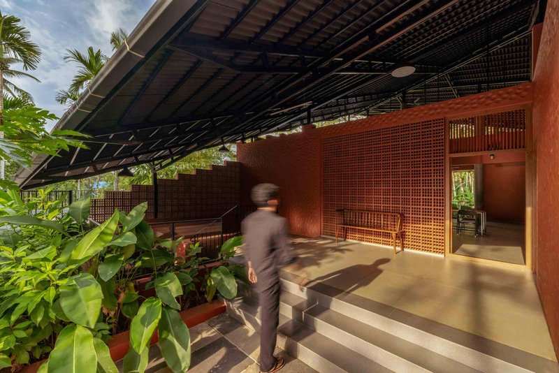 Covered outdoor walkway flanked by perforated terracotta screens and planted beds as a figure passes through sunlight