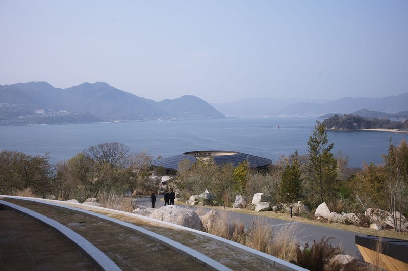 Curved roof pavilion glimpsed across a rocky planted slope with visitors walking amid native grasses