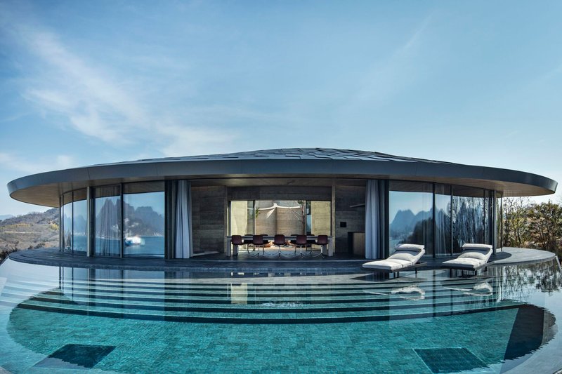 Glass-walled dining pavilion with floating roof canopy and infinity pool reflecting the mountains beyond